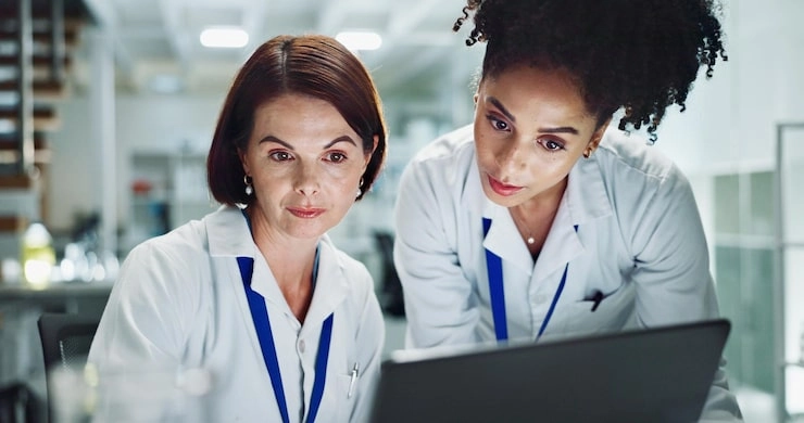 Two female scientists in lab coats (with blue lanyards) collaborating closely while reviewing data on a laptop in a modern laboratory setting