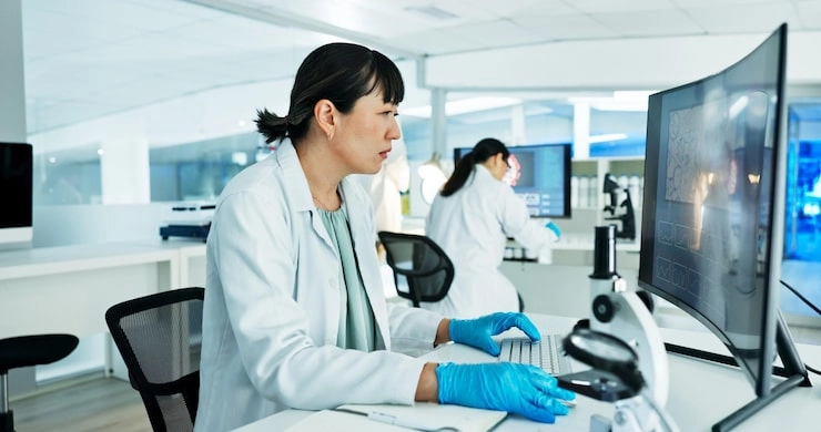 A female scientist in a white lab coat and blue gloves working at a computer in a modern laboratory, with another scientist in the background
